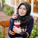 Home A woman in a hijab enjoys a refreshing drink while seated outdoors, depicting a moment of leisure.